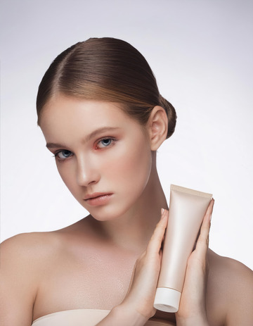 一位女性，她的头发梳成一个发髻，面容姣好，皮肤白皙，眼神明亮且富有神采。她双手捧着一个米色的护肤品软管，背景为浅色，整体画面给人一种清新、优雅且时尚的感觉