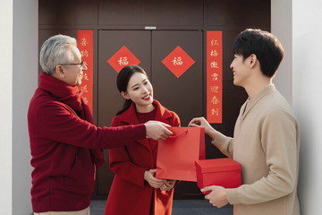 一位穿着红色衣服的老人正将一个红色购物袋递给一位同样穿着红色衣服的女子女子身旁是一位穿着米色衣服的男子手中拿着红色礼盒