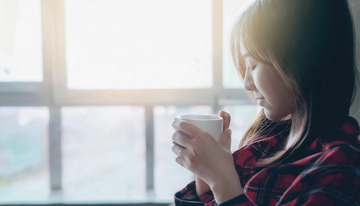 一位女子她穿着红黑格子的衣服正站在窗边双手捧着一个白色的杯子