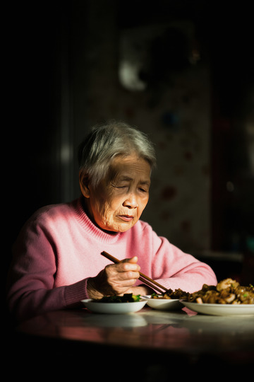一位头发花白的老奶奶她穿着粉红色的衣服独自坐在桌前用餐桌上摆放着食物老奶奶神情专注画面光线集中在她身上周围环境略显昏暗营造出一种孤独的氛围仿佛在诉说着老年人独自生活的日常1
