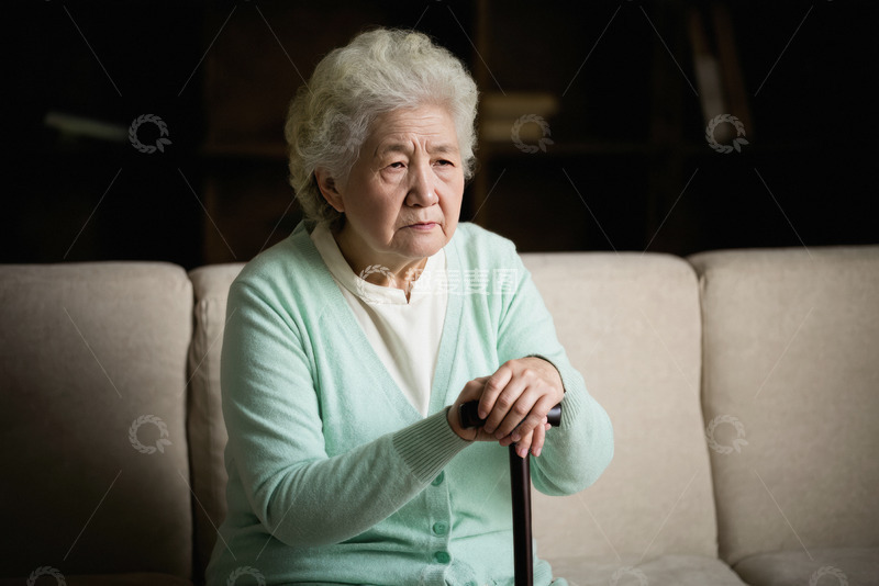 一位头发花白的老奶奶她穿着浅绿色的毛衣独自坐在沙发上双手握着拐杖神情略显落寞背景较暗突出了老奶奶的孤独感仿佛在诉说着老年人独自在家的寂寞时光