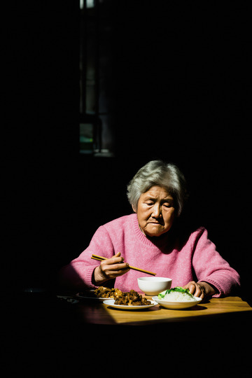 一位头发花白的老奶奶她穿着粉红色的衣服独自坐在桌前用餐桌上摆放着食物老奶奶神情专注画面光线集中在她身上周围环境略显昏暗营造出一种孤独的氛围仿佛在诉说着老年人独自生活的日常