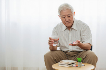 一位头发花白的老爷爷他穿着浅色的衬衫坐在室内的椅子上手里拿着药片和水杯似乎正准备服药2