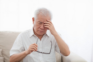 一位头发花白的老爷爷他穿着浅色的衣服坐在沙发上表情略显痛苦扶着额头似乎正被头痛困扰