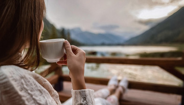 一位女子她穿着米白色的毛衣正坐在一个可以远眺山水美景的地方手中拿着一个白色的杯子似乎在悠闲地享受着眼前的自然风光
