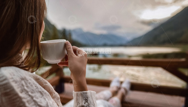 一位女子她穿着米白色的毛衣正坐在一个可以远眺山水美景的地方手中拿着一个白色的杯子似乎在悠闲地享受着眼前的自然风光