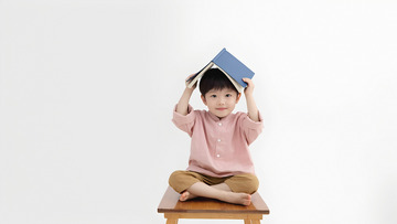 一个小男孩坐在古朴的木桌前，双手托腮，脸上带着微笑，头上戴着一顶用蓝色书本折成的帽子，显得俏皮又可爱