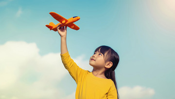 一个小女孩穿着黄色上衣，右手高高举起一架橙色的玩具飞机，仰头看向飞机，脸上带着笑容
