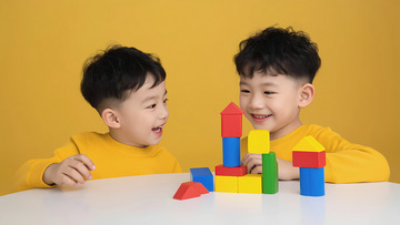 两个小男孩都穿着黄色上衣，坐在一张白色桌子前，一起玩彩色积木。他们面带笑容，看向彼此，气氛温馨愉快1