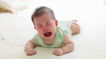 一个穿着绿白相间衣服的婴儿趴在浅色的平面上，眉头紧皱，双眼紧闭，嘴巴大张，正在哭泣，脸上露出委屈的神情。