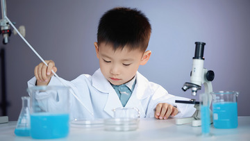 一个小男孩身着白色的实验服，专注地在桌前做实验。他右手拿着一根白色的实验棒，桌上摆放着装有蓝色液体的锥形瓶
