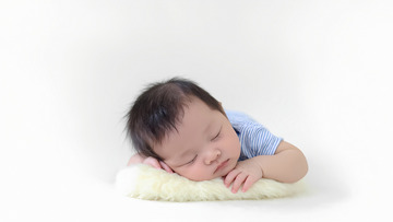 一个婴儿穿着浅蓝色的衣服，正趴在柔软的浅色毛毯上安静地睡着。婴儿的双手放在脸颊下方，头发乌黑，五官精致，表情平和，背景为纯白色，整个画面给人一种宁静，温馨的感觉