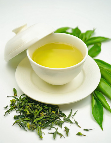 装有绿茶茶汤的白色盖碗，盖碗置于白色茶托上。盖碗的盖子部分掀开，露出黄绿的茶汤，周围散落着新鲜的绿茶叶，背景有翠绿的竹叶点缀1