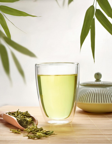 透明玻璃杯中装着泡好的绿茶，茶汤色泽黄绿明亮，杯中茶叶舒展。旁边放着装有干绿茶的茶勺，背景中有茶壶和竹叶装饰