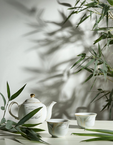 精美的白色茶具，包括茶壶和茶杯。茶具上有精致的花纹装饰，旁边摆放着翠绿的竹叶，背景是光影交错的灰色幕布，营造出一种典雅
