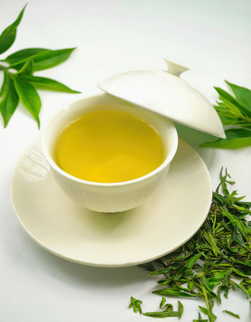 装有绿茶茶汤的白色盖碗，盖碗置于白色茶托上。盖碗的盖子部分掀开，露出黄绿的茶汤，周围散落着新鲜的绿茶叶，背景有翠绿的竹叶点缀