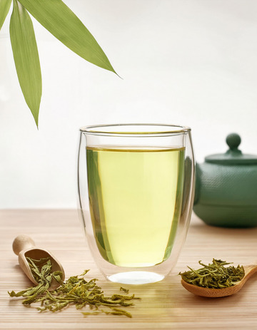 透明玻璃杯中装着泡好的绿茶，茶汤色泽黄绿明亮，杯中茶叶舒展。旁边放着装有干绿茶的茶勺，背景中有茶壶和竹叶装饰1
