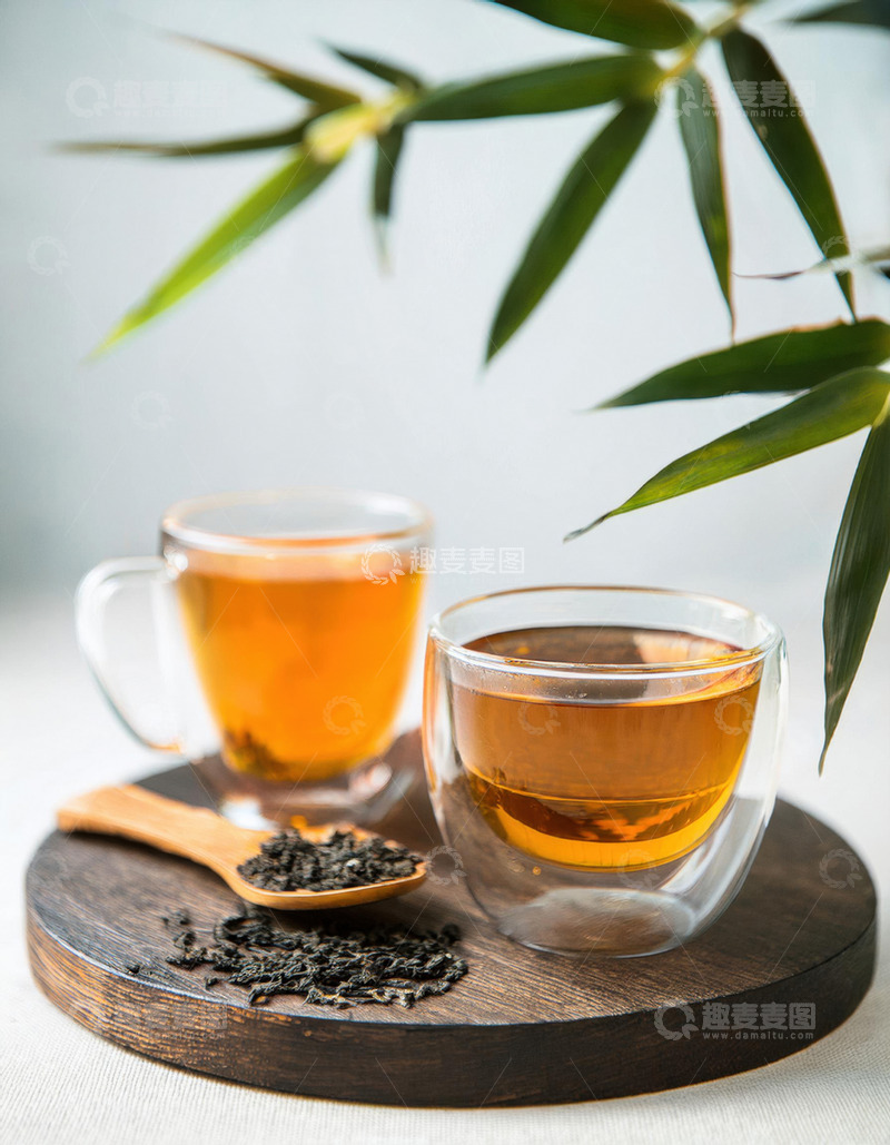高清大图下载【趣麦麦图】两个透明玻璃杯中装着橙黄色的红茶茶汤，放置在木制托盘上，旁边有装着红茶茶叶的木勺，背景中还有绿色的枝叶，整体画面清新自然