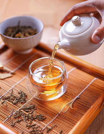 ，一只手正拿着白色茶壶往玻璃杯中倒红茶