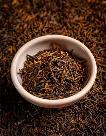 白色的碗，碗中装满了色泽乌润且带有金色芽毫的红茶茶叶，背景是大量散落的同款红茶茶叶，整体突出了红茶茶叶的质感与形态