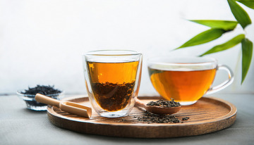 两个透明玻璃杯中装着橙黄色的红茶茶汤，放置在木制托盘上，旁边有装着红茶茶叶的木勺，背景中还有绿色的枝叶，整体画面清新自然3