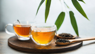 两个透明玻璃杯中装着橙黄色的红茶茶汤，放置在木制托盘上，旁边有装着红茶茶叶的木勺，背景中还有绿色的枝叶，整体画面清新自然2