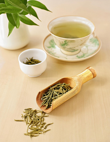 一个装满绿茶的竹制茶勺放在编织垫上，茶叶自然散落，整体画面清新雅致，旁边是白色花瓶和绿植