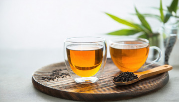 两个透明玻璃杯中装着橙黄色的红茶茶汤，放置在木制托盘上，旁边有装着红茶茶叶的木勺，背景中还有绿色的枝叶，整体画面清新自然1