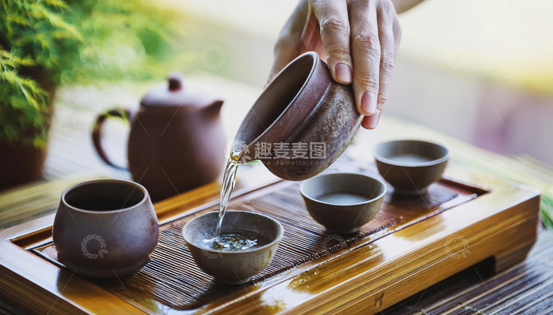 高清大图下载【趣麦麦图】-一只手握着紫砂壶，往形态古朴的茶杯中倒水，背景有绿植和其他茶具，营造出悠然的茶文化氛围