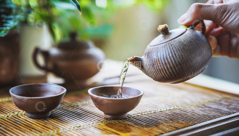 高清大图下载【趣麦麦图】-一只手握着紫砂壶，往形态古朴的茶杯中倒水，背景有绿植和其他茶具，营造出悠然的茶文化氛围2