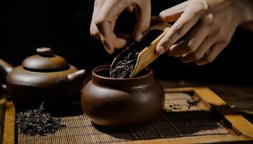 一双手正用木制茶勺将茶叶从容器舀入古朴的陶制茶罐中，背景是木质桌面和竹制茶席