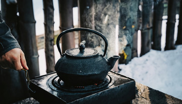 冰天雪地中，一个古朴的铁壶置于户外，壶口正冒着热气，背景是木栅栏