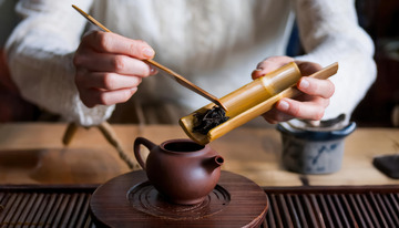 一双手正拿着竹制茶则，将茶叶倒入古朴的紫砂壶中，背景简单，展现传统的泡茶准备过程