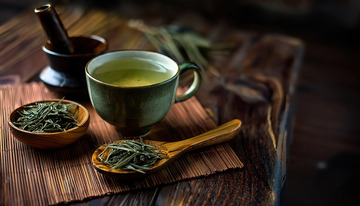 古朴的木制桌面上，放着一只盛有淡绿色茶汤的茶杯，旁边有装满茶叶的木勺和木碗，后方还有一个捣具，整体营造出浓郁的茶文化氛围