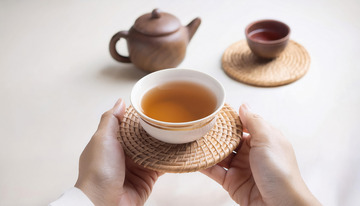 一双手捧着置于编织杯垫上的茶杯，杯中盛有橙黄色茶汤，背景有古朴的茶壶和茶具