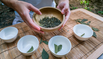一双手捧着装有茶叶的木碗，面前的竹制垫子上摆放着几只白色小茶杯，部分茶杯中放置着茶叶，背景似乎是户外自然环境，营造出一种闲适的茶文化氛围