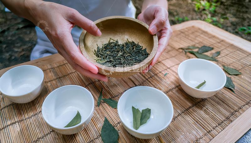 高清大图下载【趣麦麦图】一双手捧着装有茶叶的木碗，面前的竹制垫子上摆放着几只白色小茶杯，部分茶杯中放置着茶叶，背景似乎是户外自然环境，营造出一种闲适的茶文化氛围
