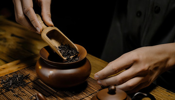 一双手正用木制茶勺将茶叶从容器舀入古朴的陶制茶罐中，背景是木质桌面和竹制茶席1