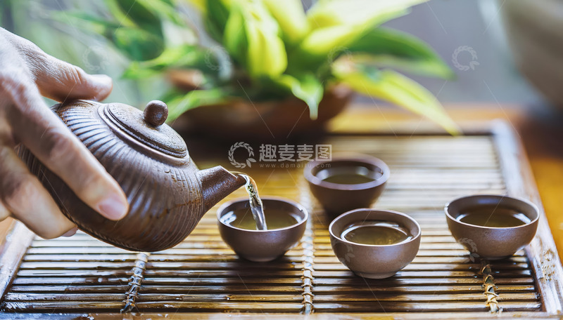 高清大图下载【趣麦麦图】-一只手握着紫砂壶，往形态古朴的茶杯中倒水，背景有绿植和其他茶具，营造出悠然的茶文化氛围1