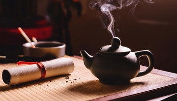 一把古朴的黑色茶壶置于竹制茶席上，壶口冒着袅袅热气，旁边放着一卷绑着红绳的卷轴和一个装有茶勺等物品的容器