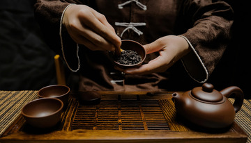 一双手穿着中式服饰，正从容器中抓取茶叶放入茶杯，旁边摆放着古朴的紫砂壶和其他茶具，背景较暗，突出主体，营造出传统的茶文化氛围
