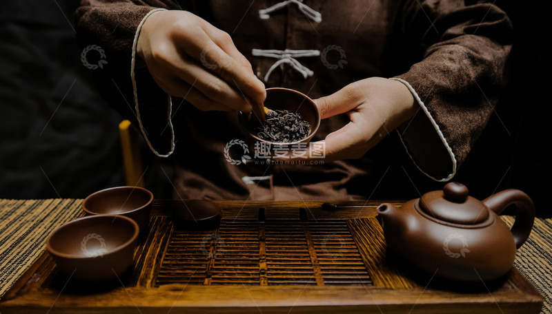 高清大图下载【趣麦麦图】一双手穿着中式服饰，正从容器中抓取茶叶放入茶杯，旁边摆放着古朴的紫砂壶和其他茶具，背景较暗，突出主体，营造出传统的茶文化氛围