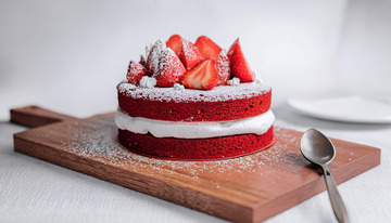 一块放在木制砧板上夹着白色奶油顶部有草莓装饰撒着糖粉的红色蛋糕旁边有银勺