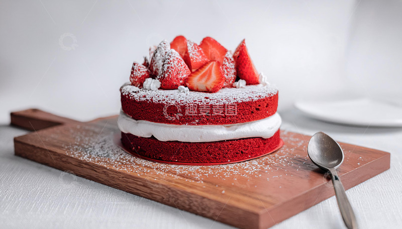 高清大图下载【趣麦麦图】-一块放在木制砧板上夹着白色奶油顶部有草莓装饰撒着糖粉的红色蛋糕旁边有银勺