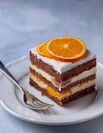 一块放在白色盘子上夹着奶油和橙子片顶部有巧克力片装饰的蛋糕旁边有把叉子2