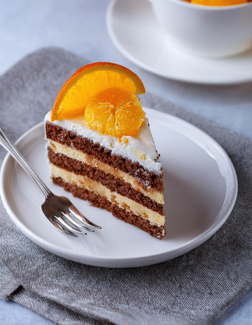 一块放在白色盘子上夹着奶油和橙子片顶部有巧克力片装饰的蛋糕旁边有把叉子1