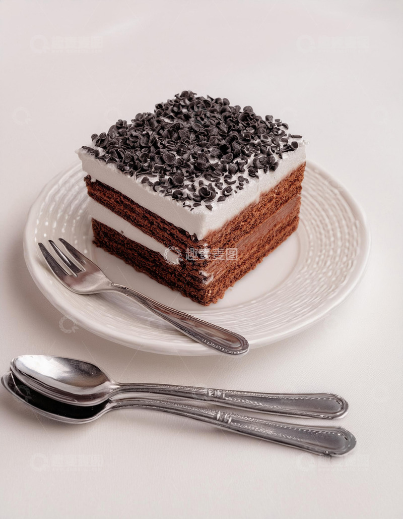 一块放在白色盘子上夹着奶油顶部撒有巧克力屑的方形蛋糕旁边有把银勺和银叉1