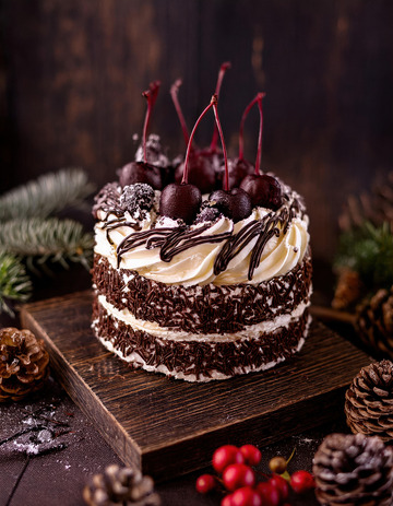 放在深色盘子上装饰有樱桃奶油和巧克力屑周围有松枝松果的巧克力蛋糕1