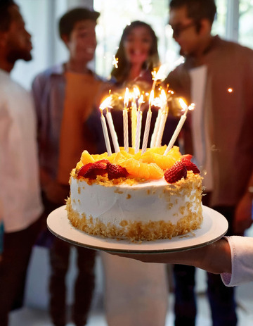 一只手端着一个装饰有水果插着点燃蜡烛的生日蛋糕背景中有几个人影