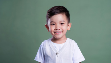 小男孩穿着白色背心和灰色短袖面带笑容站在绿色背景前的图片